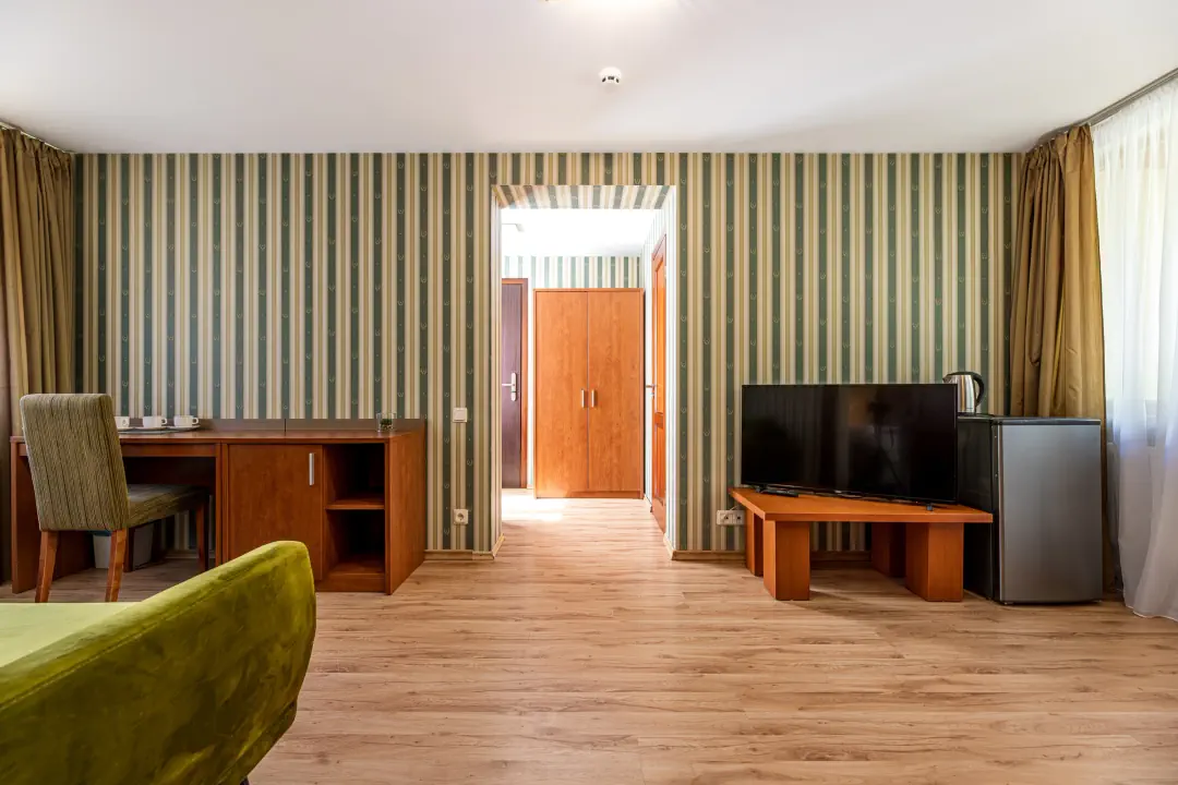 Numerio svetainė: žalia sofa, stalas su kėde, televizorius ant žemos spintelės, šaldytuvas ir durys į koridorių