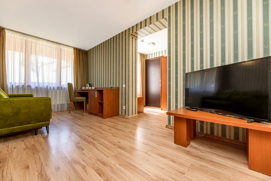 Svetainė: didelis televizorius ant medinės spintelės, stalas su kėde prie lango, žalia sofa