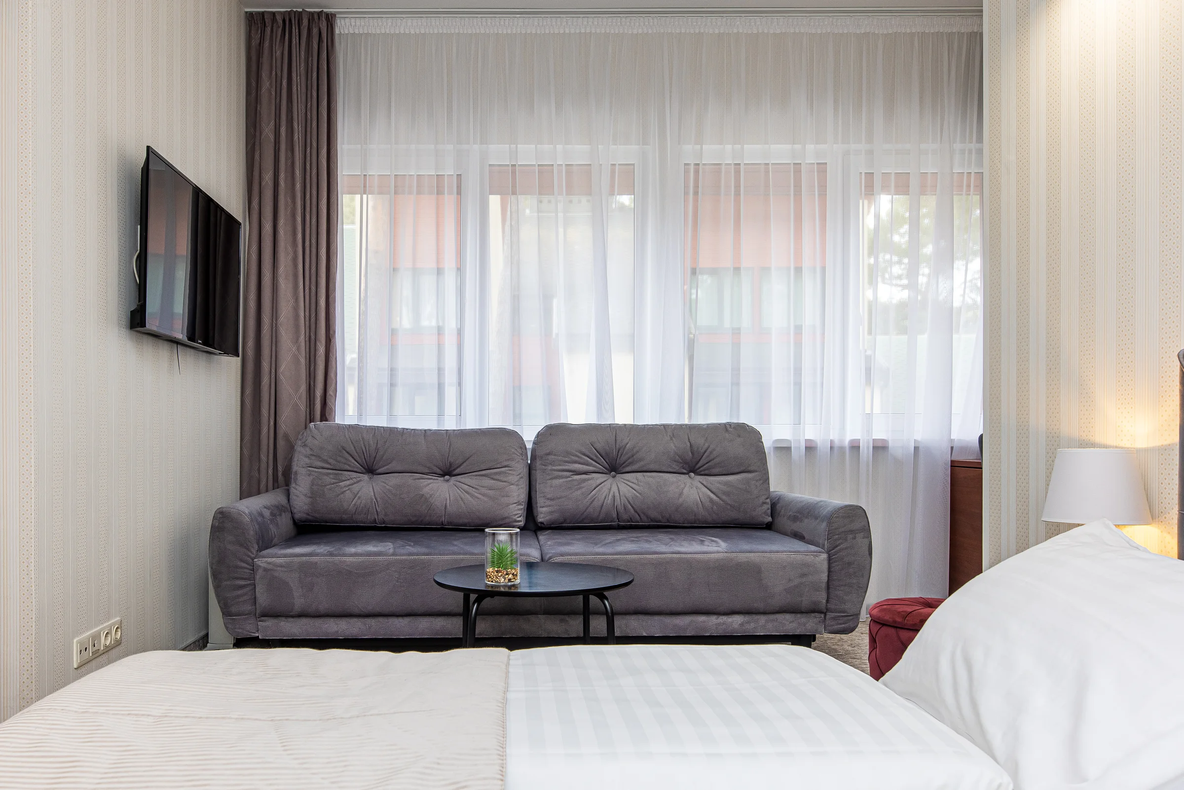 Televizorius ant sienos, pilka sofa po langu ir lovos kojūgalis