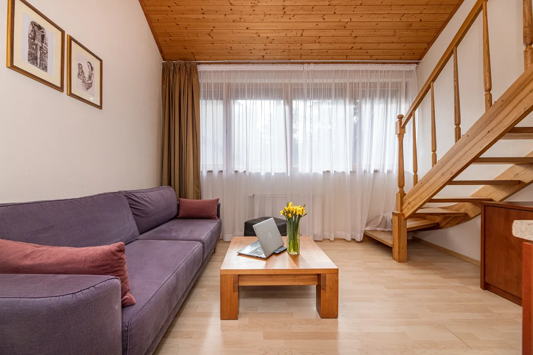 Apartamentų svetainė pirmame aukšte: minkštas kampas, kavos staliukas, langas ir mediniai laiptai į miegamuosius